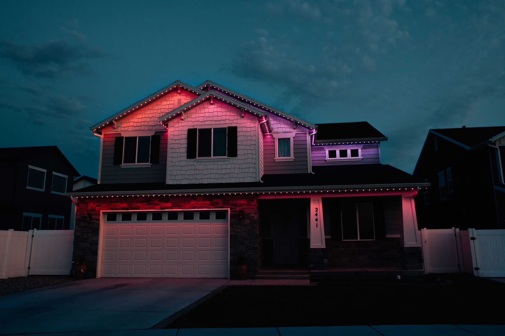 A two-story home with red and purple permanent roofline lighting under a cloudy dusk sky