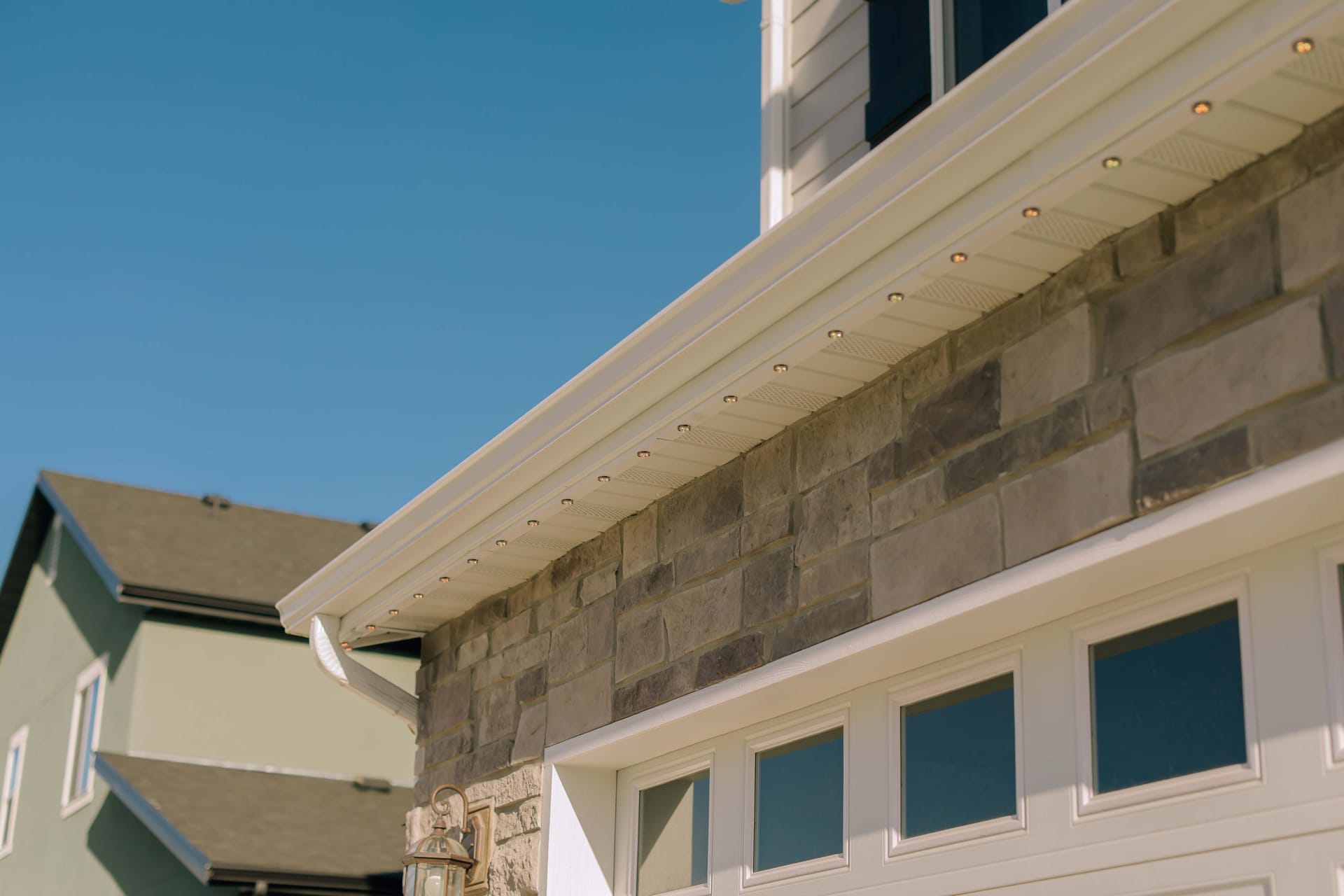 Discreet recessed permanent lighting installed in a row along a home's white soffit and eave over gray stone veneer